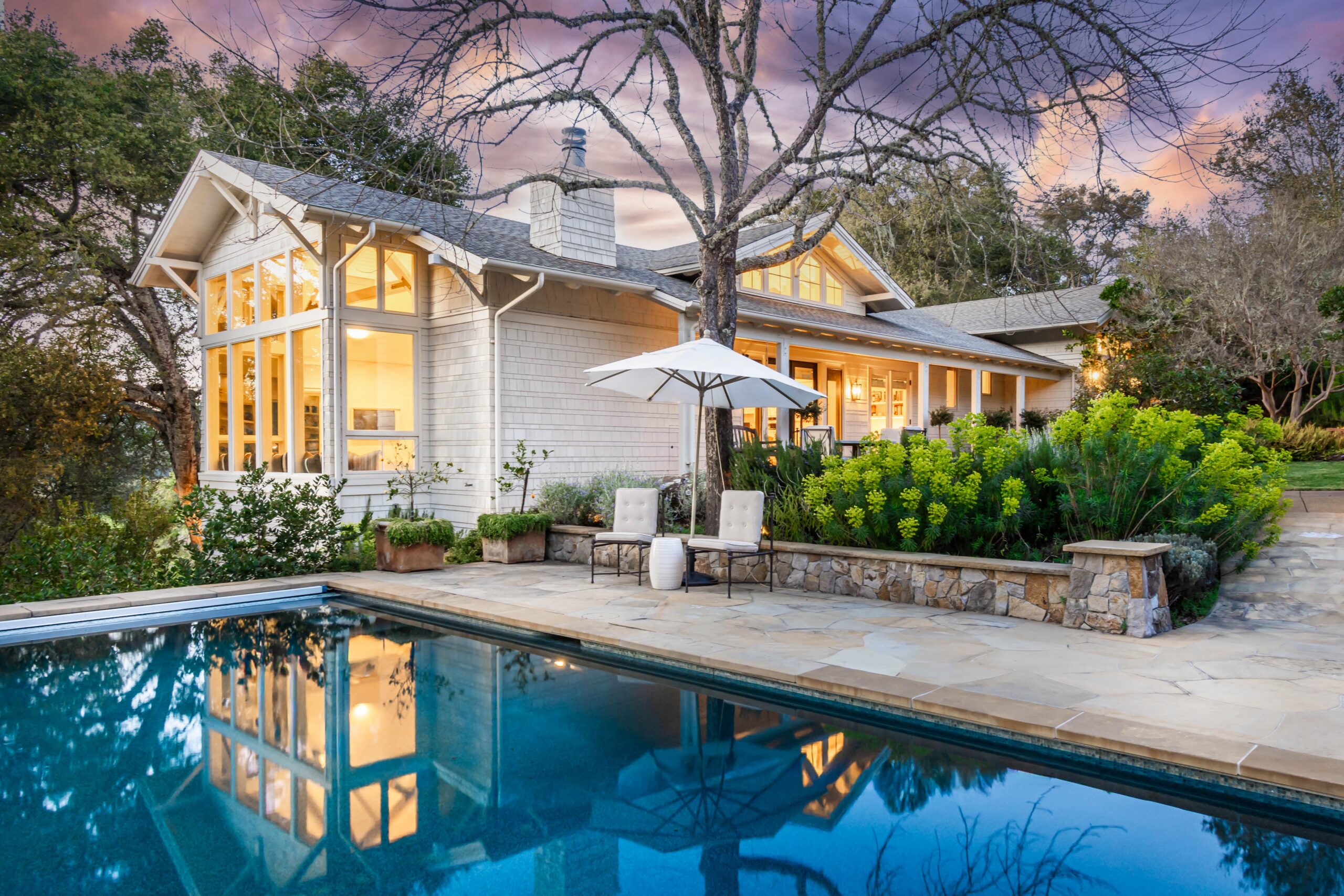 A modern house with large windows and warm lighting is surrounded by trees and lush landscaping. In the foreground, a swimming pool, stone patio, two chairs, and an umbrella await under a colorful May 2026 sunset—an A-list retreat.