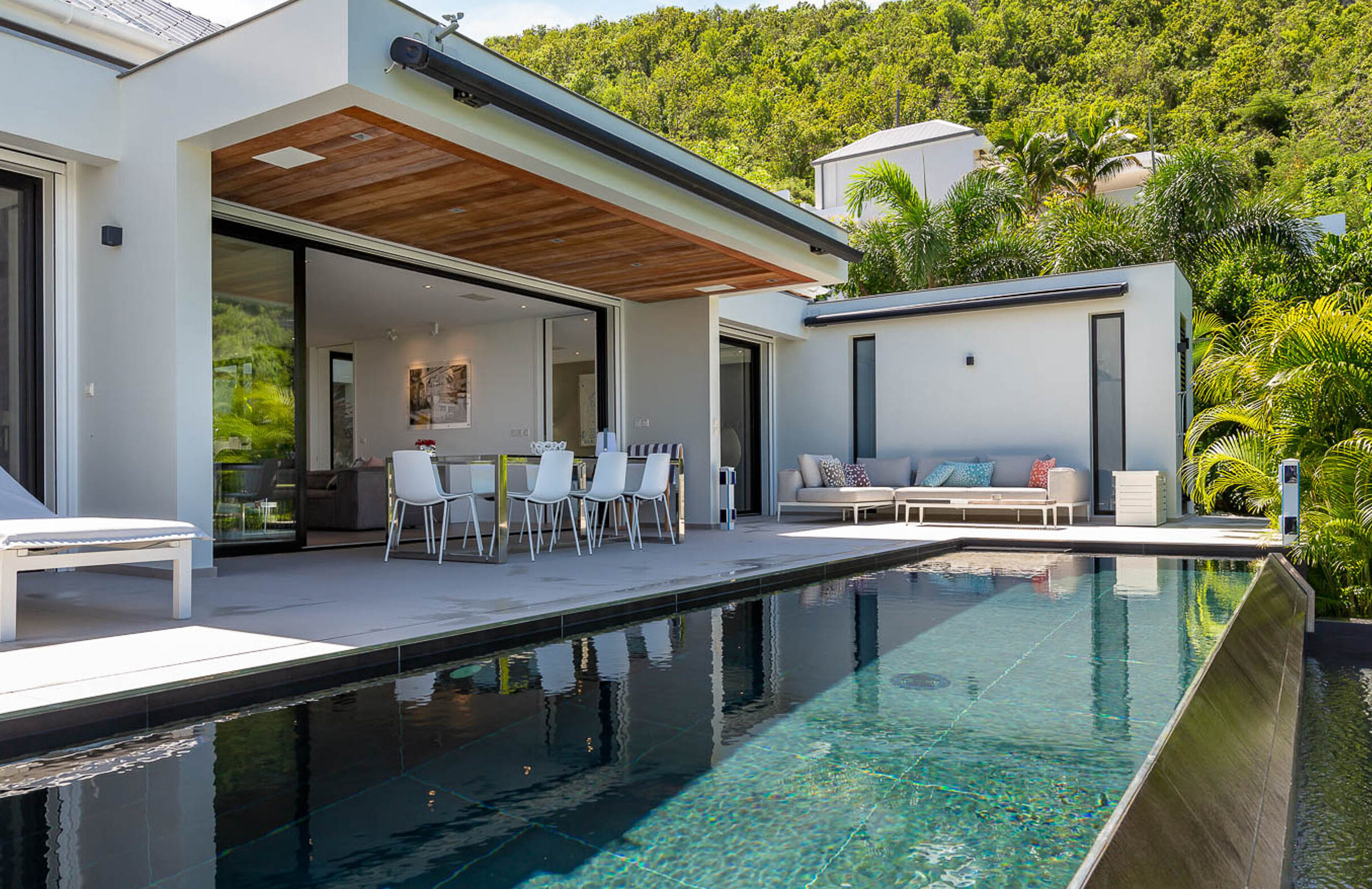 Modern villa with a shaded outdoor dining area, lounge seating, and a sleek rectangular swimming pool. Featured in A-list: April 2026, the space is bright, open, and inviting for relaxation amid lush greenery and hills.
