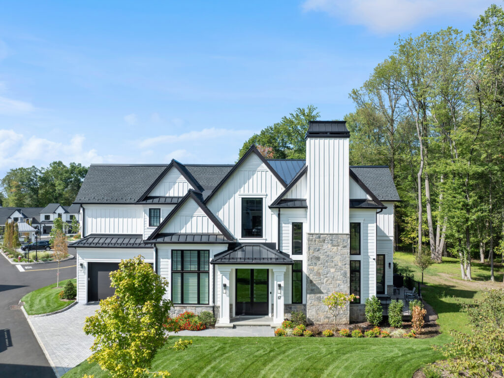 A modern two-story house with white vertical siding, dark trim, and a tall stone chimney overlooks a golf course, surrounded by a green lawn, landscaped plants, and trees under a bright blue sky.