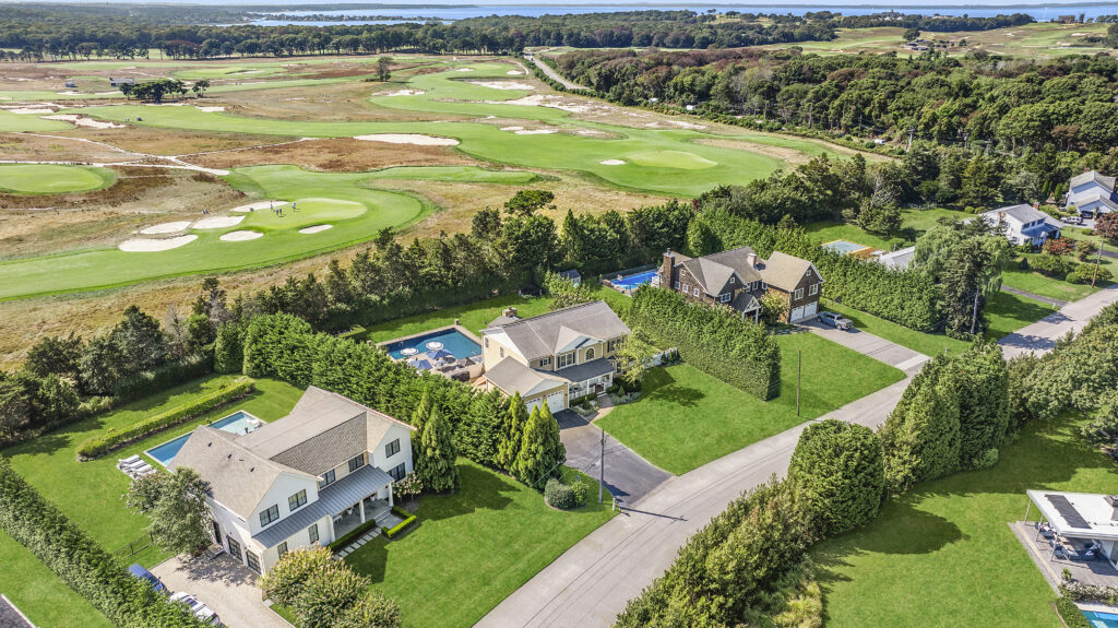 Aerial view of a suburban neighborhood with large houses, green lawns, private pools, tree-lined streets, and a scenic golf course in the background near a tranquil water body and wooded areas.