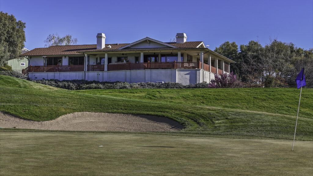 A scenic golf course green with a sand bunker in front, a blue flag on the hole, and a large clubhouse featuring a red railing and covered porch on a hill under a clear blue sky.
