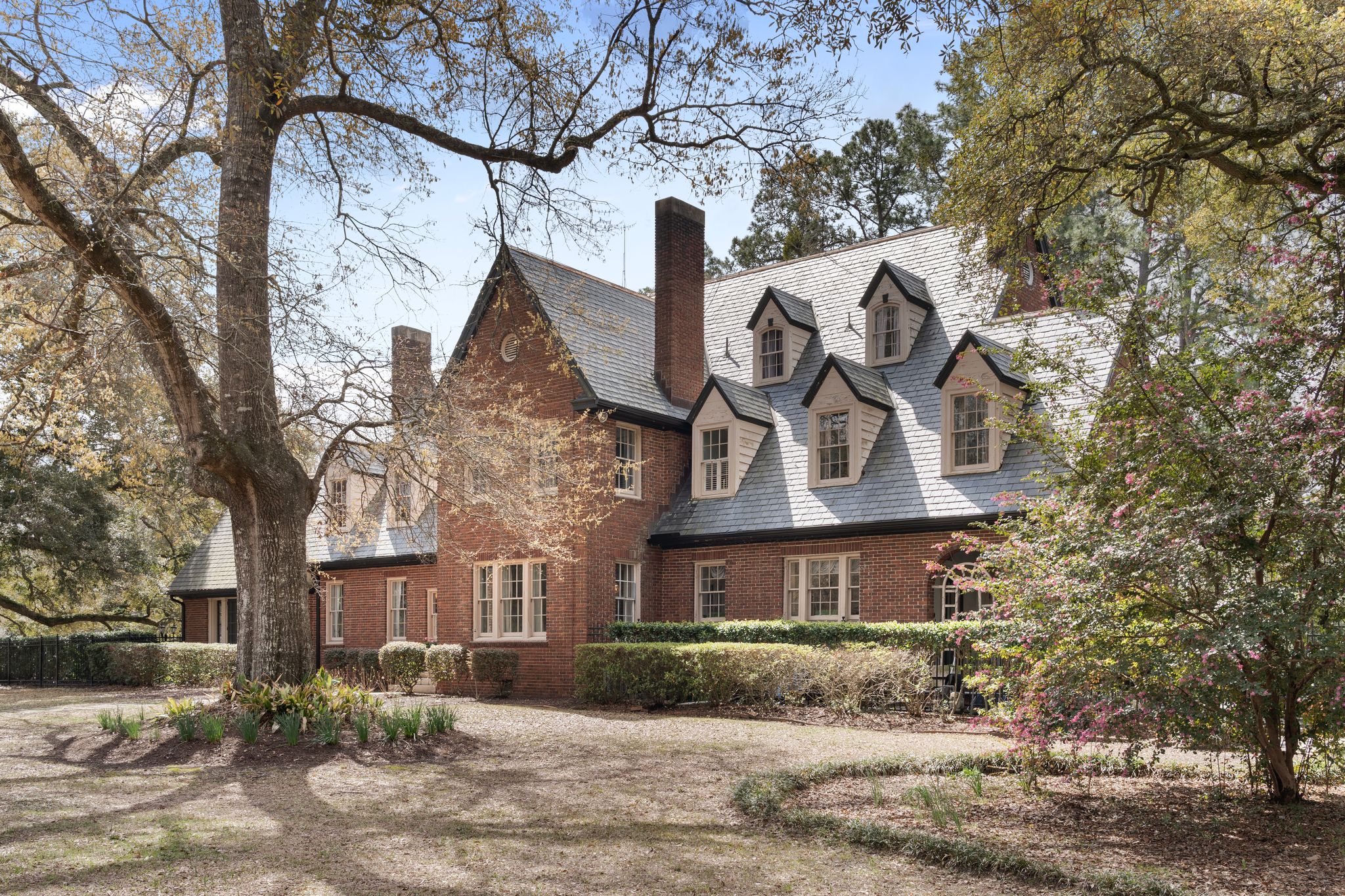 A large brick house with steep, gabled roofs and multiple chimneys, surrounded by tall trees and landscaped bushes, stands out like an a-list residence on a sunny May 2026 day.