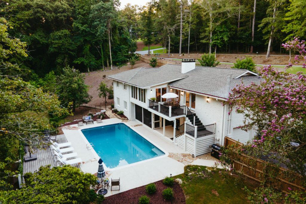 A large white house with a spacious backyard is perfect for enjoying Taurus season, featuring an outdoor swimming pool, lounge chairs, a deck, patio seating, and surrounding lush trees.