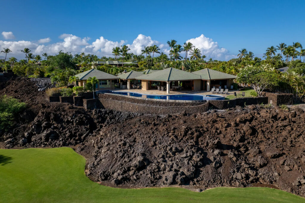 A luxury house with green roofs sits on a rocky lava field, surrounded by palm trees and lush greenery, with a swimming pool in front and a well-manicured lawn overlooking a nearby golf course under a blue sky with clouds.