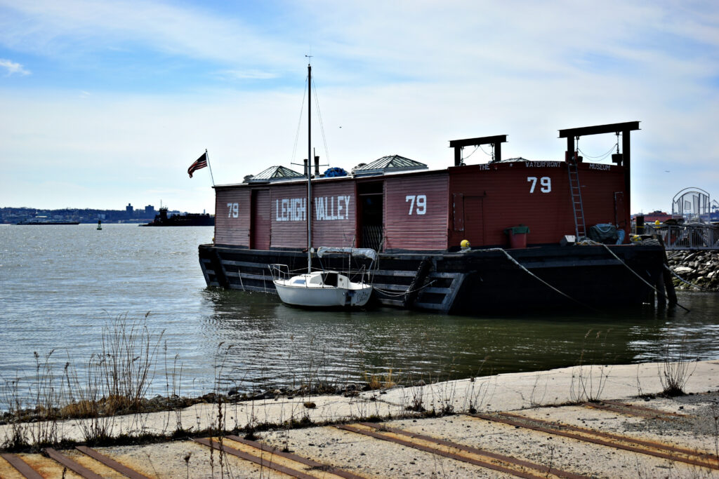 Lehigh Valley Railroad Barge No. 79 A small white sailboat is docked next to the large red barge Lehigh Valley 79—one of the area's ordinary landmarks—on calm water, with an American flag flying and a distant shoreline visible under a cloudy sky.