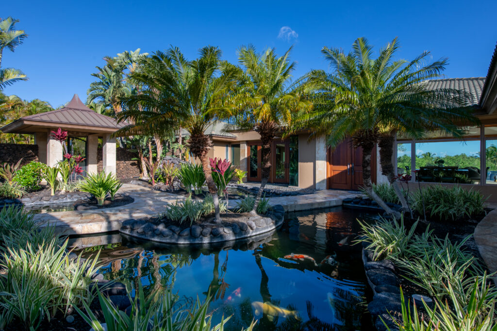 A tropical garden with palm trees and lush plants surrounds a koi pond with clear water and colorful fish, creating a serene retreat perfect for relaxing during Taurus season, next to a modern house with large windows and a covered walkway.