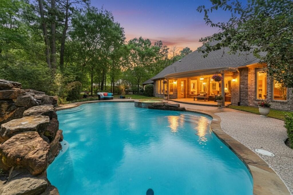 A backyard pool with rock waterfall feature, surrounded by trees and greenery, next to a brick house glowing at dusk—perfect for unwinding during Taurus season. Patio furniture and a hot tub invite relaxation in this tranquil setting.