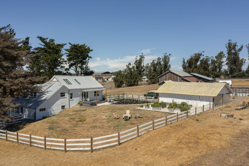 A white farmhouse with a metal roof sits next to a shed, both enclosed by a white wooden fence on dry, brown land—an idyllic scene often found in equestrian estates—with scattered trees and distant barns under a clear blue sky.
