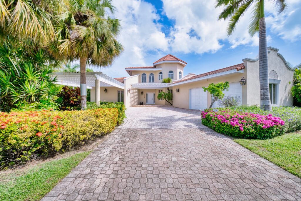 A large, two-story Mediterranean-style house with arched windows, a tiled roof, and a wide driveway lined with palm trees and colorful landscaping, perfect for relaxing during Taurus season on a bright, partly cloudy day.
