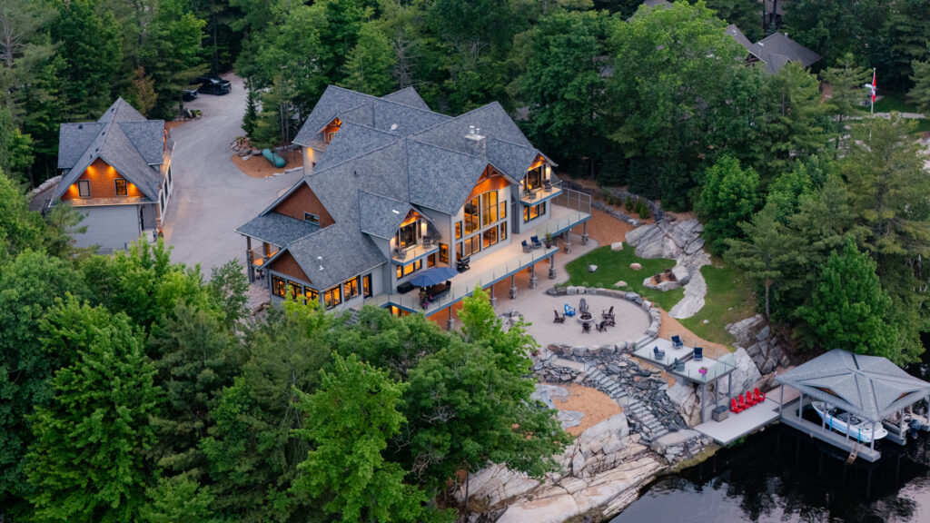 Aerial view of a large, modern lakeside house surrounded by trees, with a stone patio, fire pit, gazebo, and dock. Several people gather outside near the water, soaking in the tranquil vibes of Taurus season.