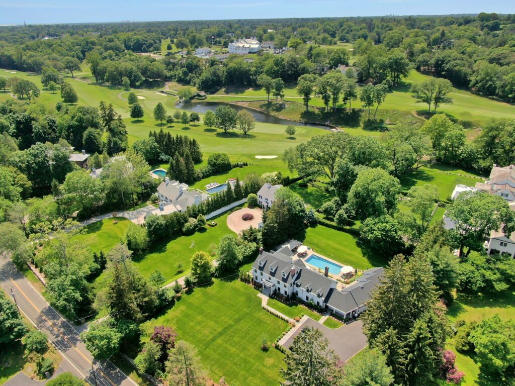 Aerial view of a lush, green suburban neighborhood basking in Taurus season, with large houses, manicured lawns, swimming pools, winding roads, and a golf course dotted with ponds and trees in the background.