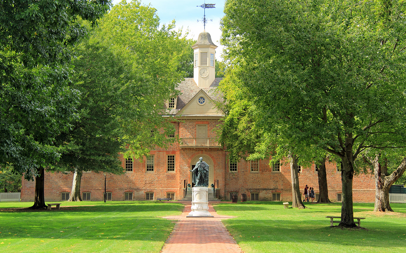 A brick colonial-style building with a clock tower and cupola is surrounded by green trees. A statue stands on a brick path leading to the entrance, with grass and benches on either side.