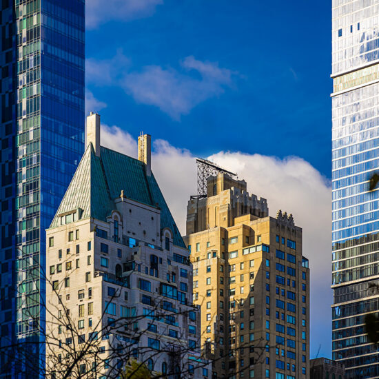 Several high-rise buildings of varying architectural styles stand close together against a vibrant blue sky with clouds, framed by tree branches in the foreground—an iconic view in the heart of Manhattan luxury sales.