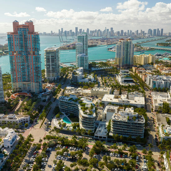 Aerial view of South Florida real estate with tall modern buildings, turquoise water, palm trees, and a distant skyline under a partly cloudy sky.