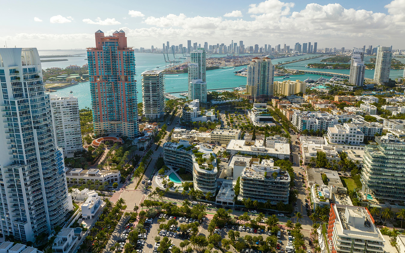 Aerial view of South Florida real estate with tall modern buildings, turquoise water, palm trees, and a distant skyline under a partly cloudy sky.