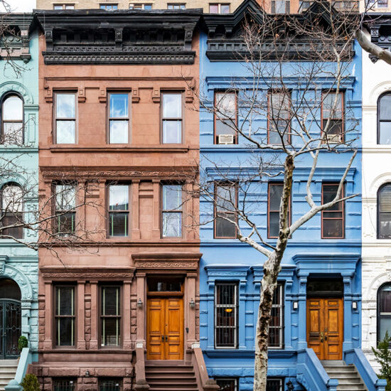 Four colorful Brooklyn condo brownstone townhouses in a row, painted green, brown, blue, and white, with bare trees in front and classic architectural details, each featuring stoops and wooden front doors.