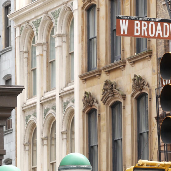 A close-up of a street sign reading W Broadway and a yellow traffic light, set against historic stone buildings with arched windows—capturing the charm of the Manhattan real estate market in an urban setting.