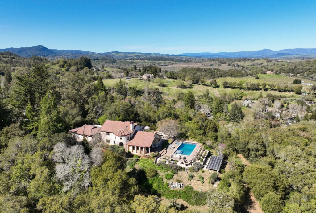 A large villa with a red-tiled roof sits among trees, featuring a swimming pool, garden, and solar panels. Set within equestrian estates, the property overlooks rolling hills, distant mountains, and clear blue skies.