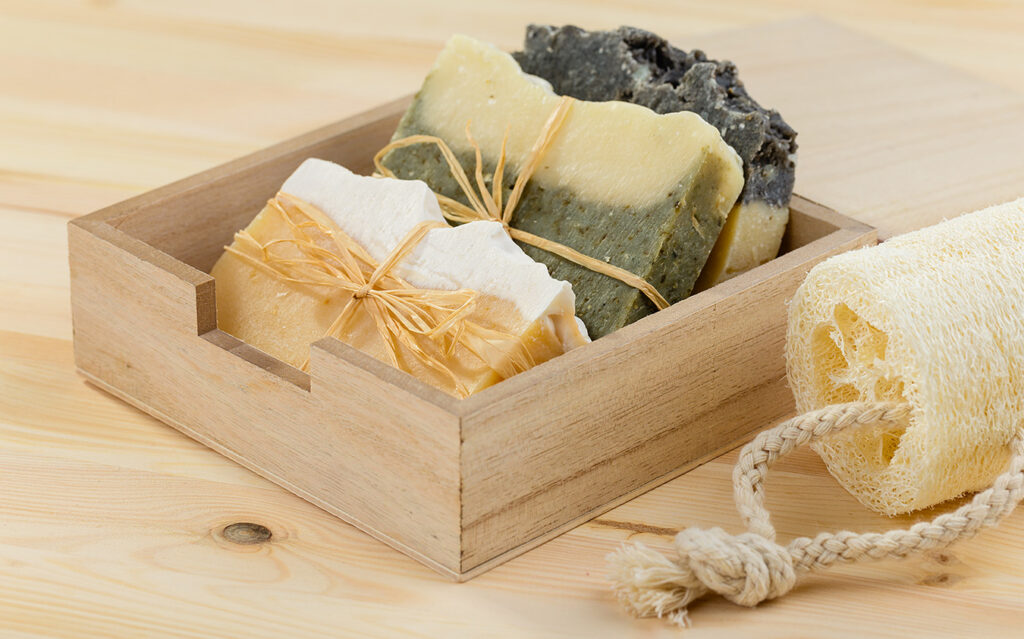 A wooden box containing three handmade soap bars tied with raffia, next to a natural loofah sponge with a rope handle, ideal for holiday decorating, all placed on a light wooden surface.