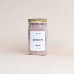 A clear glass jar with a gold lid filled with pink bath salts. The white label reads SERENITY and Lavender Scented Bath Soak, making it a perfect addition to your bathroom or as a touch of holiday decorating against a neutral beige background.