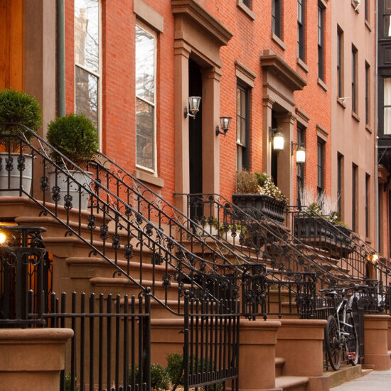 A row of classic brownstone townhouses with black iron railings and neatly trimmed shrubs lines a quiet city street, bathed in warm daylight.