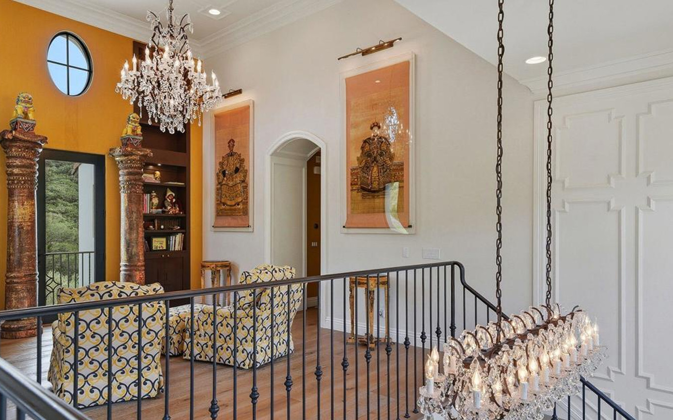 A bright, elegant upstairs sitting area with two patterned armchairs, chandeliers, large framed art, decorative columns, bookshelves, and a black railing overlooking the space below. Warm lighting and yellow accent wall are visible.