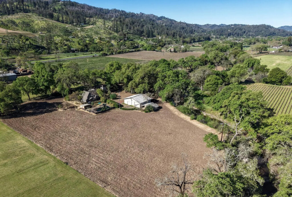 Aerial view of a rural landscape with a farmhouse, barn, trees, and tilled field surrounded by green hills, vineyards, equestrian estates, and scattered homes under a clear blue sky.