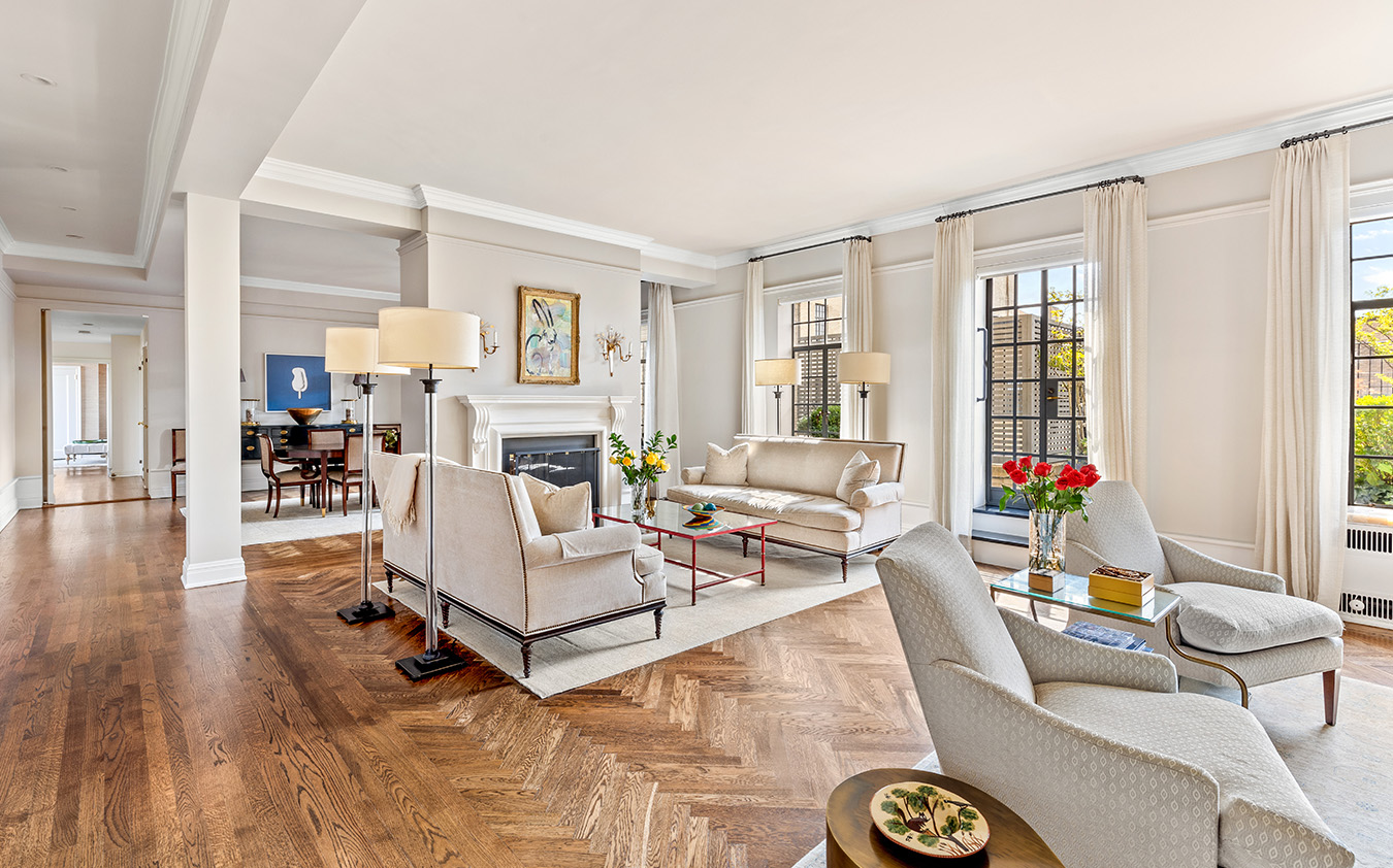 Bright, elegant living room with cream sofas, armchairs, a fireplace, hardwood floors, large windows, and tasteful decor including vases of flowers and a coffee table with a tray of snacks. Dining area visible in the background.