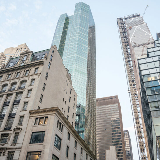 A street-level view of several tall buildings in a city, featuring a mix of older stone architecture and modern glass skyscrapers—perfectly capturing the vibrant atmosphere surrounding a Manhattan condo under a bright, partly cloudy sky.
