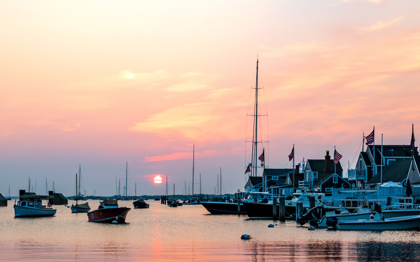 Sailboats and motorboats docked by quaint houses with flags at a harbor during a colorful sunset, with the sun low on the horizon and a sky of pink and orange hues reflected on the calm water.
