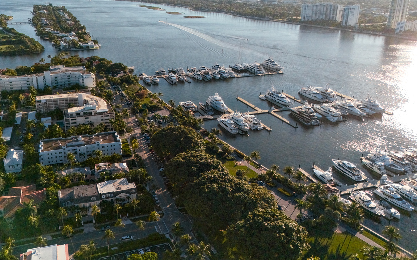 Aerial view of a marina filled with yachts and boats docked along the waterfront, surrounded by South Florida real estate, palm trees, and residential areas, with shimmering water extending into the distance.