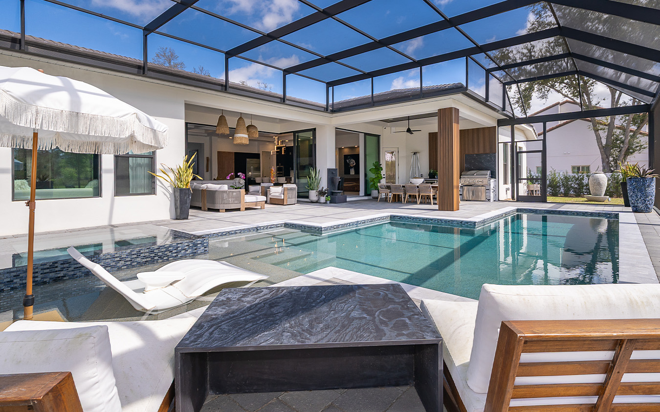 Modern outdoor patio with a swimming pool, lounge chairs, umbrella, and covered seating area. Glass walls and ceiling enclose the space, offering an a-list: May 2026 vibe with stylish interiors visible through open sliding doors.