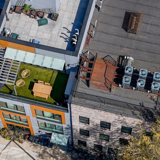 Aerial view of two adjacent rooftops in an NYC residential rental; one features a green terrace with a picnic table and chairs, while the other has several HVAC units and metal structures. Trees and sidewalks are visible at street level.