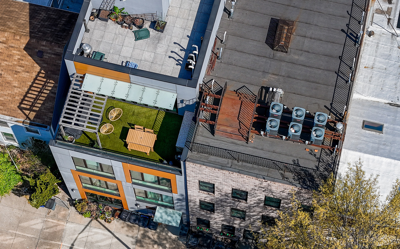 Aerial view of two adjacent rooftops in an NYC residential rental; one features a green terrace with a picnic table and chairs, while the other has several HVAC units and metal structures. Trees and sidewalks are visible at street level.
