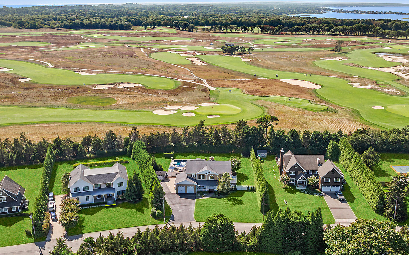Aerial view of three large houses with green lawns along a tree-lined street, adjacent to an expansive golf course featuring sand traps and winding paths, with water visible in the distance.