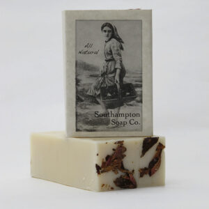 A bar of soap with dried rose petals on top sits in front of a packaged soap labeled Southampton Soap Co., perfect for holiday decorating, featuring a vintage-style image of a woman carrying buckets and the words All Natural.