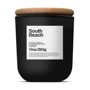 A black candle jar with a cork lid, labeled South Beach in bold text. Featuring all-natural, hand-poured coconut wax and a wood wick, this 10oz/283g candle is perfect for holiday decorating.