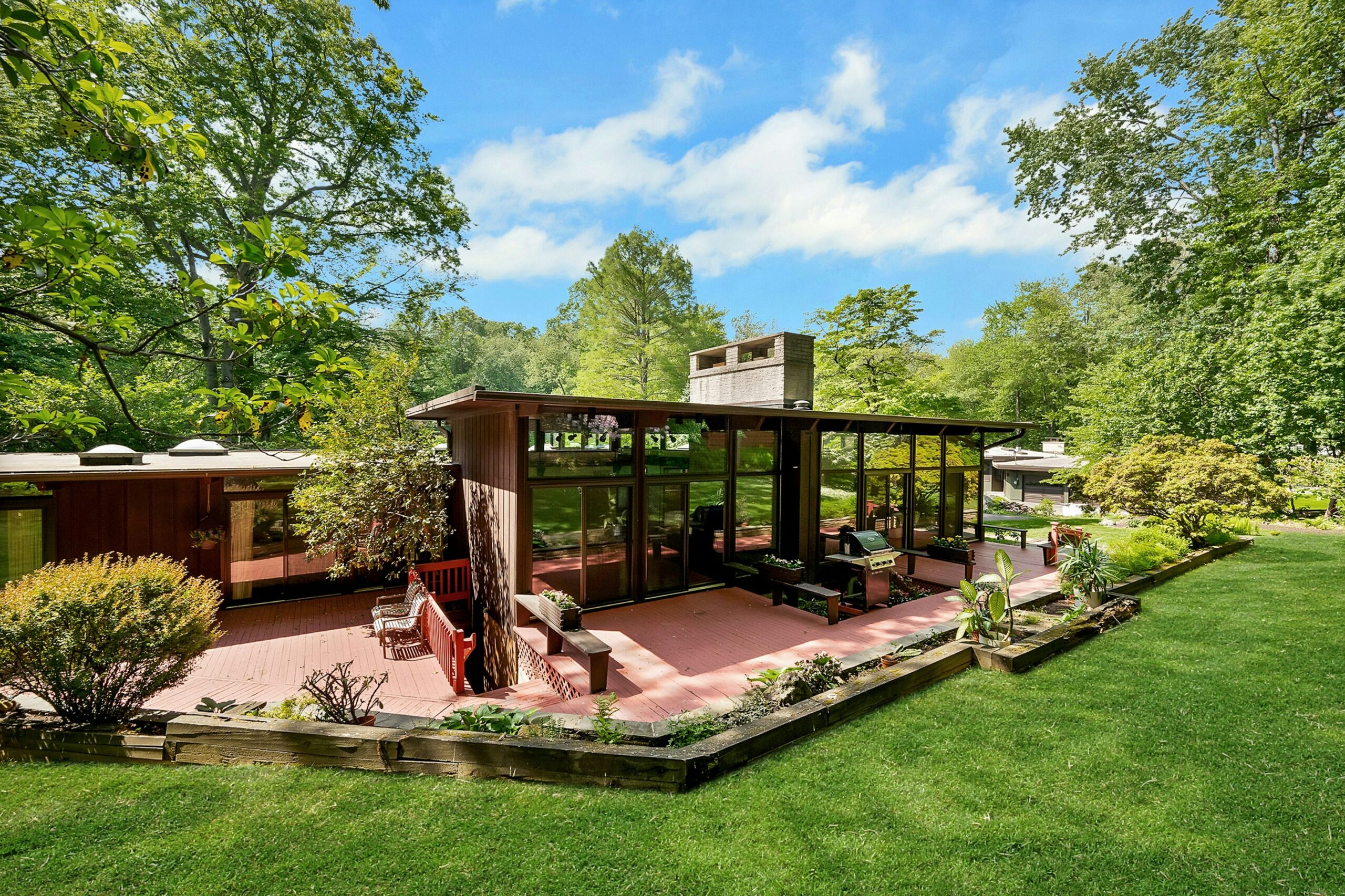 032-493003-MC8_6720_14645638 A mid-century modern house with large floor-to-ceiling windows, surrounded by a spacious patio and lush green lawn, nestled among tall trees under a bright blue sky—an A-list retreat for June 2026.