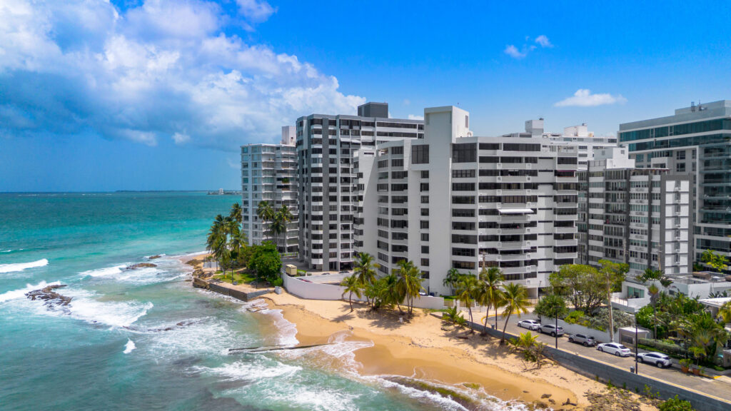 High-rise white waterside homes line a sandy beach with palm trees, where gentle waves from turquoise ocean water meet the shore under a bright blue sky with scattered clouds.