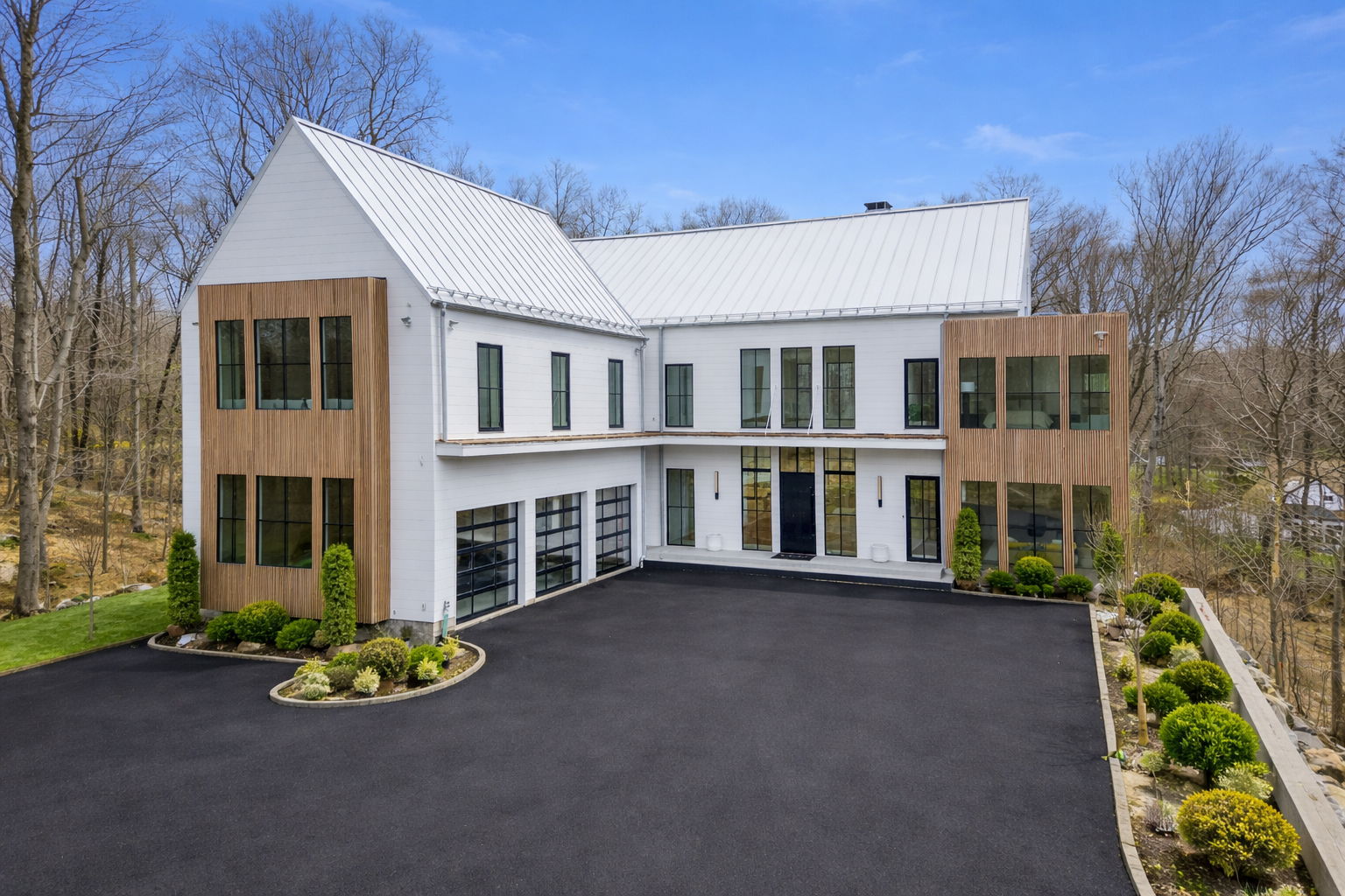 1 Modern two-story house with white walls, large windows, and wood accents on the exterior, featured on the a-list: June 2026, surrounded by landscaped shrubs and a black paved driveway, with bare trees in the background under a clear blue sky.