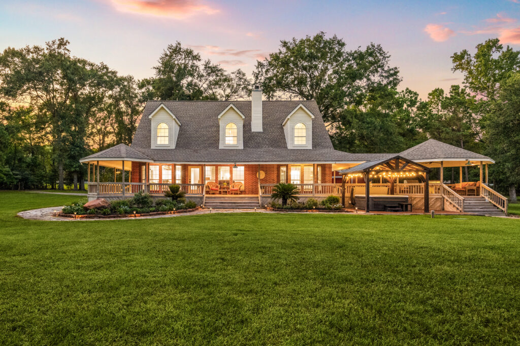 A large, two-story brick house with white trim features a wide front porch, wraparound deck, and gazebo, set against tall trees at sunset, with a well-manicured green lawn in the foreground.