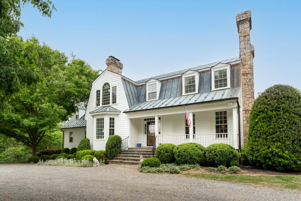 White two-story house with gray metal roof, stone chimneys, and a covered front porch. The front steps lead to a gravel driveway, landscaped with green bushes and trees. An American flag hangs by the entrance.