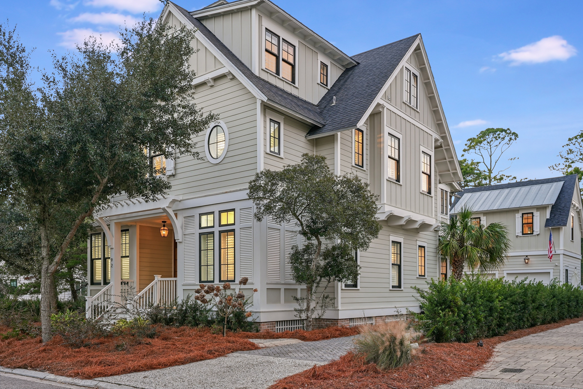 1_92 coopersmith ln - awp media - 001 A two-story light gray house with white trim, large windows, and a covered front porch is surrounded by trees, shrubs, and a gravel driveway under a blue sky—a-list: June 2026.