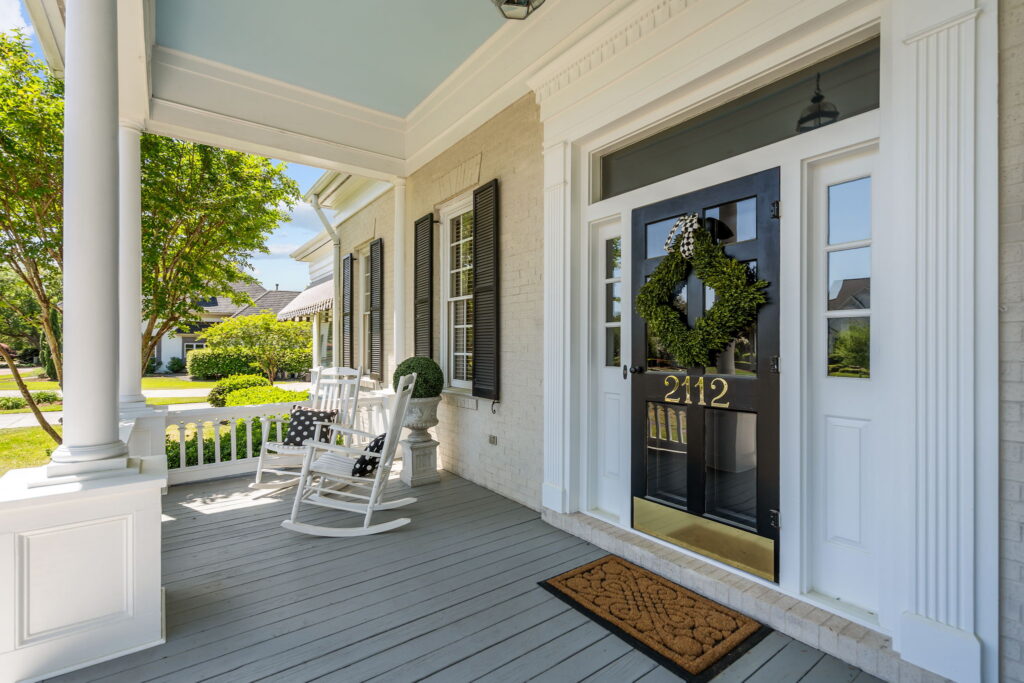Inviting front porch with white columns, a black front door displaying 2112, a green wreath, white rocking chairs with cushions, a small shrub in a pot, and a doormat on light gray wooden flooring.