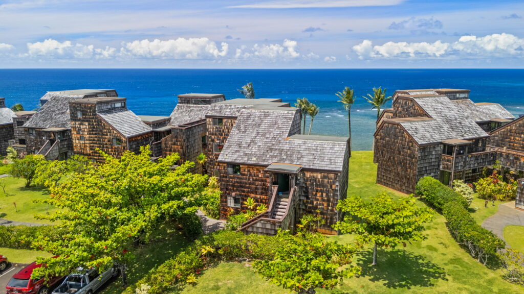 Aerial view of rustic wooden waterside homes with shingle roofs, surrounded by green trees and grass, overlooking a blue ocean under a partly cloudy sky.
