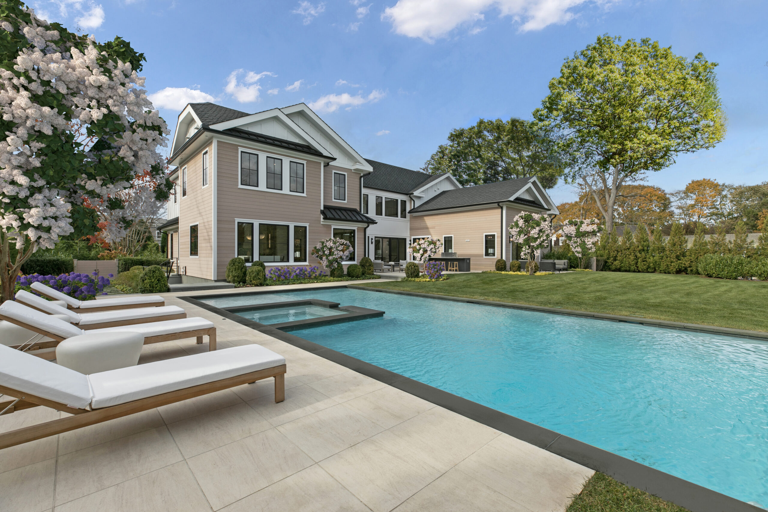 3M4A2740 A modern house with large windows and beige siding sits beside a rectangular swimming pool, ready for an a-list June 2026 gathering. Lounge chairs dot the green lawn amid trees and flowering bushes under a blue sky with white clouds.