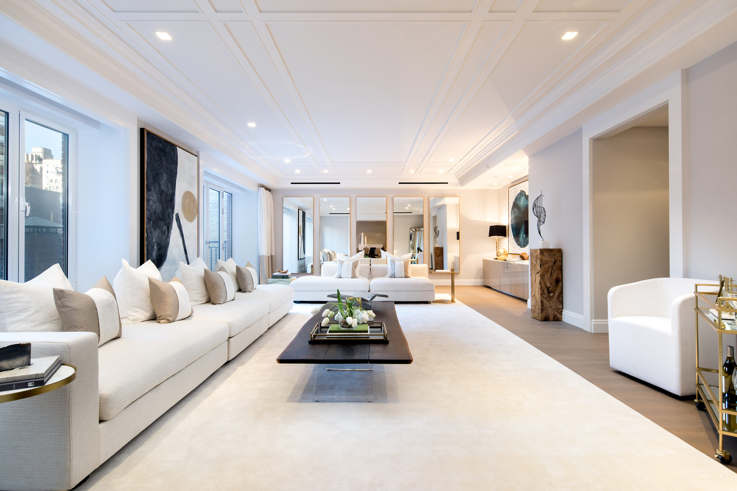 40_E_72_PH__NField-1 A spacious, modern living room with large cream sofas, a dark wood coffee table, contemporary art on the walls, floor-to-ceiling windows, and mirrored panels reflecting soft natural light—styled for an A-list: June 2026 ambiance.