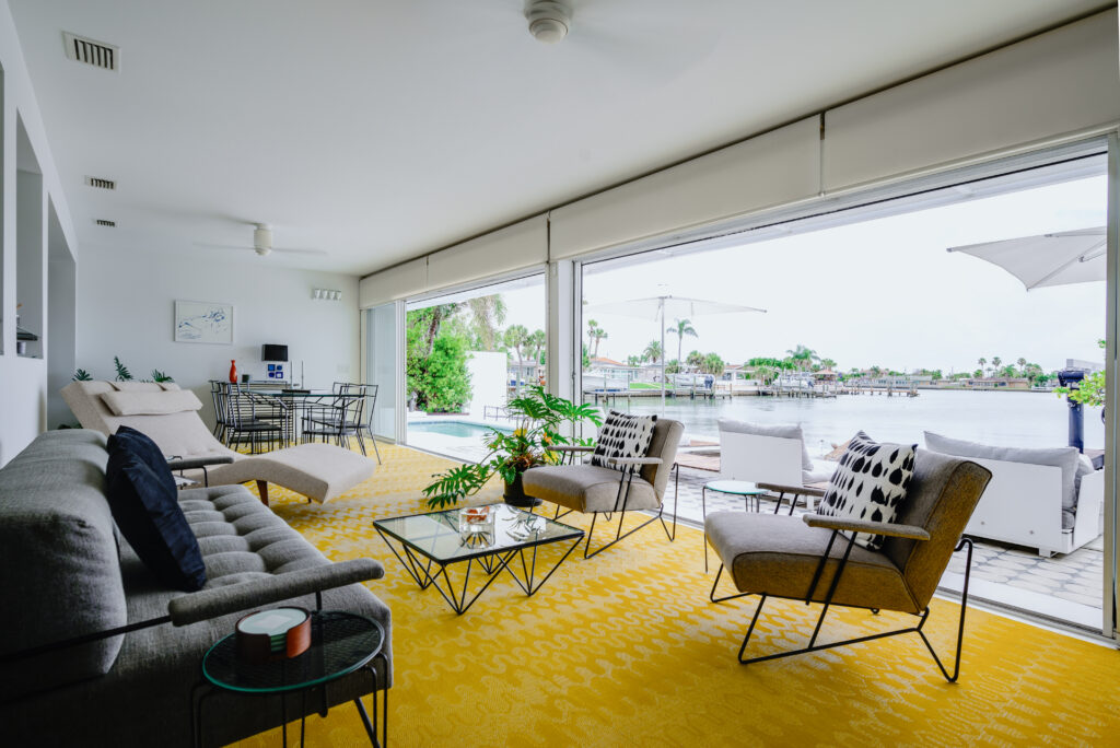 Modern living room with large glass doors opening to a waterfront patio. This inviting space in waterside homes features gray sofas, armchairs, a yellow patterned rug, glass coffee tables, and views of water, palm trees, and docks outside.
