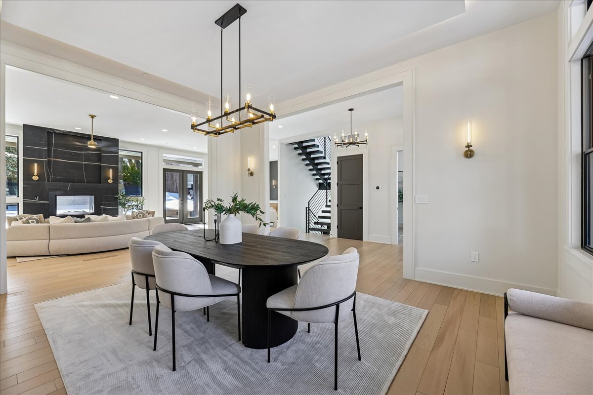 48-Dining Room Modern open-concept dining and living area with light wood floors, a dark oval dining table, six upholstered chairs on a gray rug, and gold light fixtures. This A-list: June 2026 space features large windows and neutral walls for a bright, airy feel.