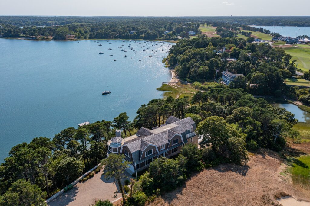 Aerial view of a large house surrounded by trees near the waterfront, with boats scattered on the water and waterside homes and lush greenery lining the distant shoreline.
